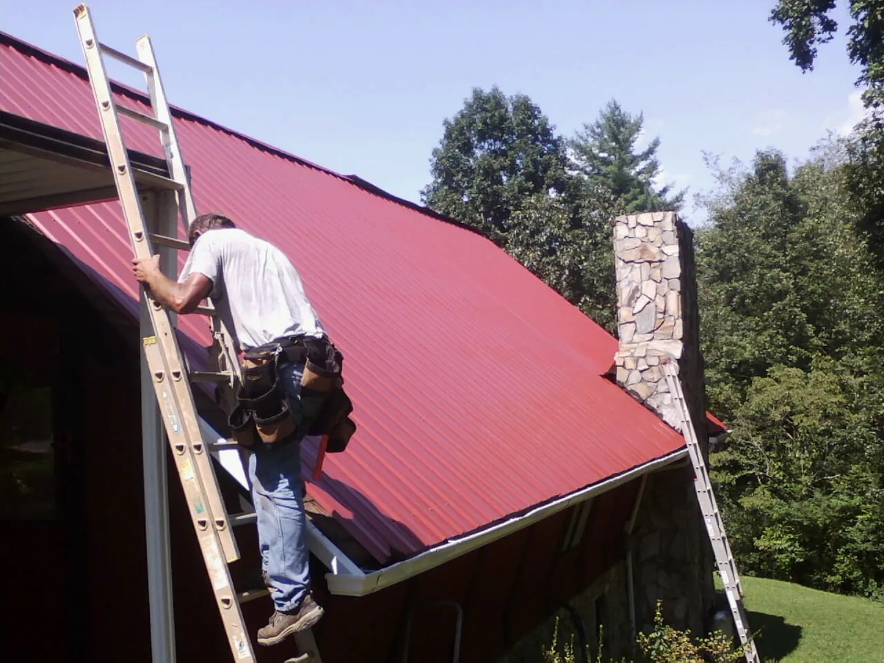 Roofer on a ladder beside a red metal roof and stone chimney — Soffit & Fascia Repair by Roofing Brothers in Cherokee