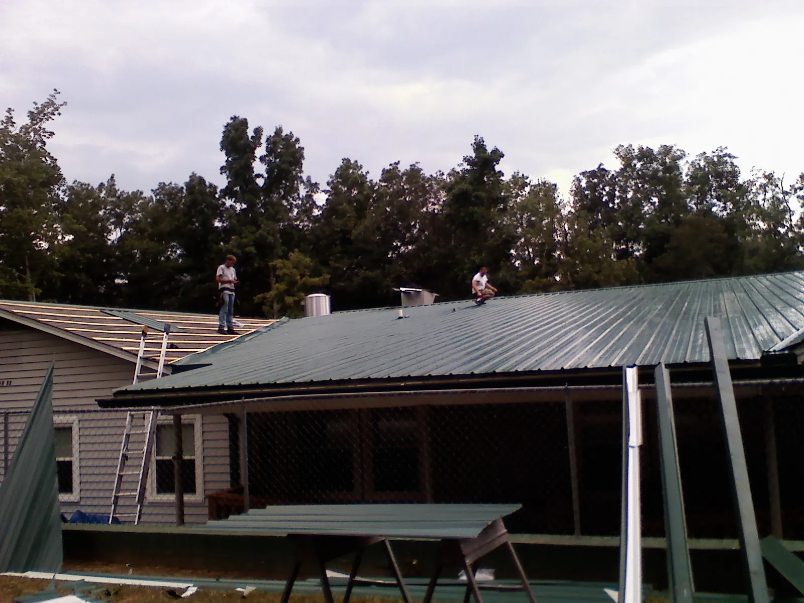 Two roofers installing green metal panels above a covered porch in Cherokee